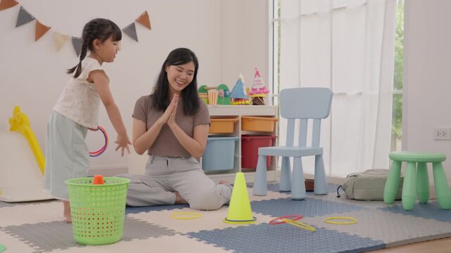 Asian mother and daughter play colorful ring toss indoors with child toys, toy basket, chair, foam mat, traffic cone, and bright window light, creating joyful family bonding in cozy home playroom