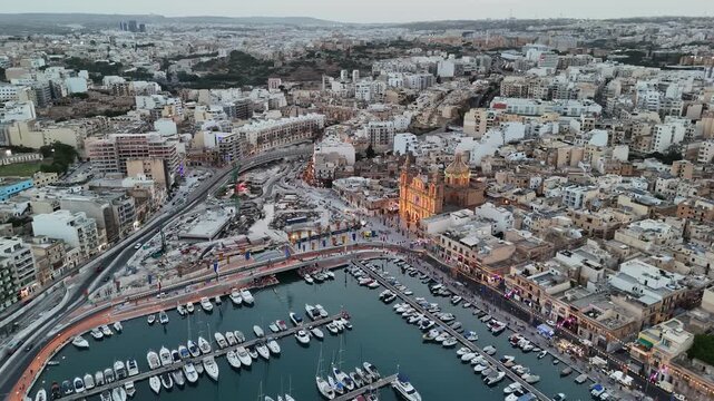Msida, Malta, Knisja Parrokkjali tal-Imsida and marina illuminated during a village festa, with boats, waterfront streets, crowds and city lights in an aerial panoramic view.