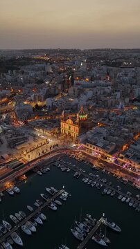 Msida, Malta Knisja Parrokkjali tal-Imsida and marina with festive lights, crowds and boats along the waterfront at night, vertical drone view of the harbor skyline.