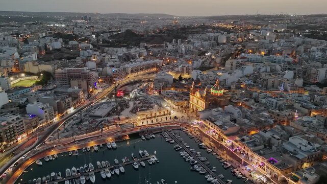 Msida, Malta village festa around Knisja Parrokkjali tal-Imsida beside the marina and harbor, with illuminated waterfront, boats and apartments at dusk, filmed by drone.