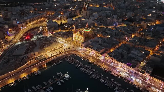 Msida marina in Malta with boats, yachts and illuminated waterfront buildings at night, featuring a coastal cityscape, church and harbor roads in an aerial view.