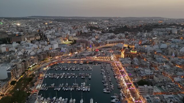 Msida, Malta waterfront and Knisja Parrokkjali tal-Imsida during a village festa at dusk, with crowds, boats and illuminated Mediterranean cityscape in an aerial view.