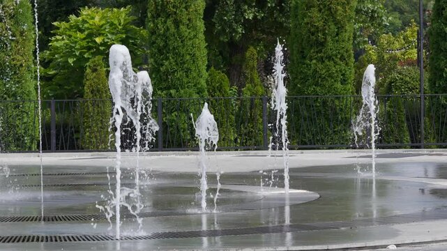 Close-up of a dry deck pedestrian fountain splashing water streams in a city park in 4K slow motion. Water jets shooting up from the ground tiles against green thujas and trees background