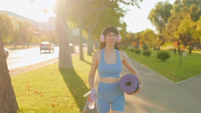 Front view of fit female walking with a yoga mat in the sunny summer park.