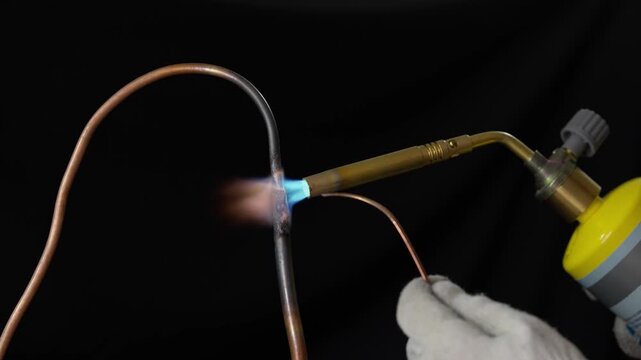 Close-up of a plumber brazing copper pipe with a propane gas torch in 4K slow motion. High temperature soldering process of metal tubing with a hot blue flame against a black background, industrial HV