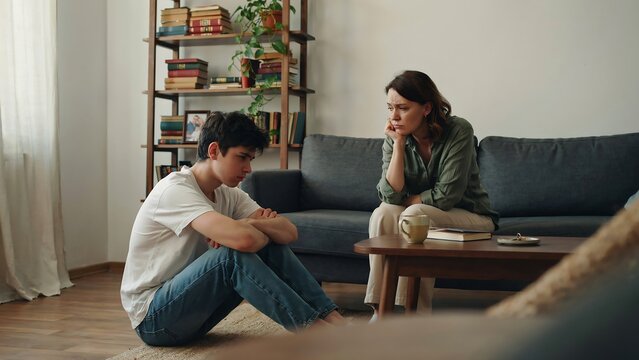 Worried mother sitting on couch looking at her unhappy teenage son who is sitting on the floor feeling very sad