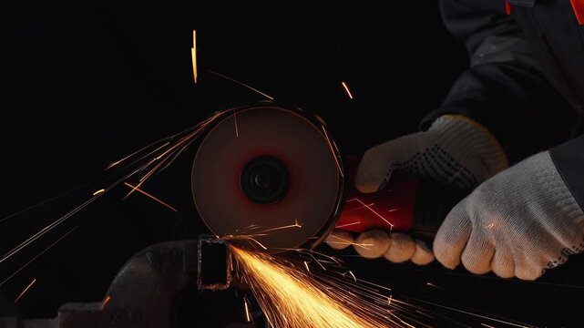 Close-up of a worker in protective gloves cutting a metal square pipe with an angle grinder in 4K slow motion. Stream of bright orange sparks flying from the abrasive disc against a dark background