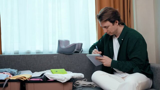 Man packing his suitcase for summer vacation travel, writing in a notebook to check his packing list. Baggage preparation and tourism concept. High quality 4k footage