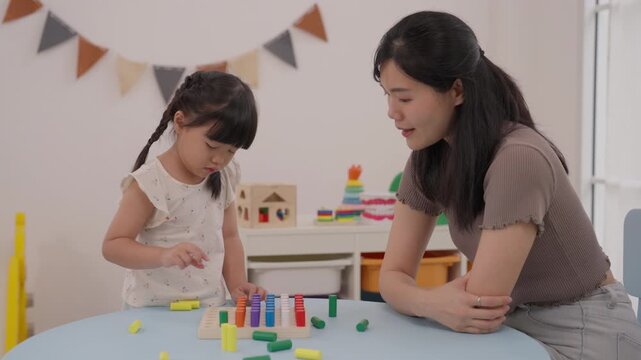 Mother Teaching Child Counting Colorful Blocks During Playful Home Learning Activity, Asian Mother and Daughter Building Wooden Toy Together at Table in Bright Indoor Educational Setting