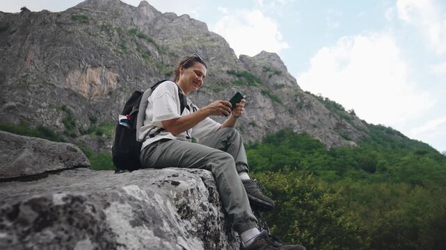 Young female traveler with a backpack using a smartphone while sitting on a large rock with a scenic mountain view during a hiking adventure.