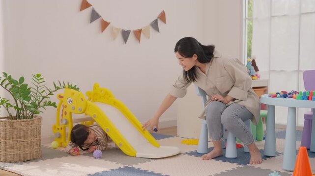 Asian mother and toddler playroom activity, family bonding indoors as parent helps child enjoy colorful slide and toy play, creating joyful home learning moment with warm care and natural window light