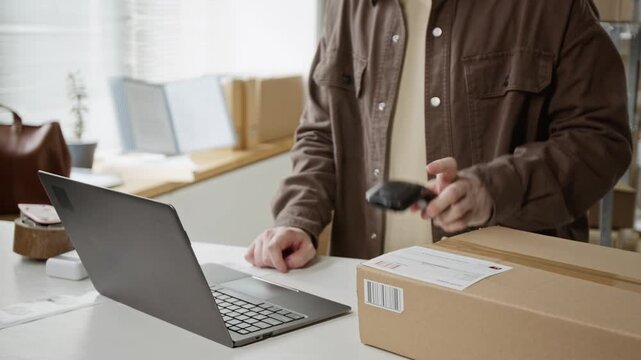 Caucasian young adult male typing on laptop and scanning barcode on cardboard box in warehouse. Clip is ideal for e-commerce, small business shipping, and logistics marketing videos