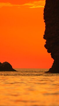 Golden sunset illumination reflects off ocean waves surrounding jagged volcanic rocks near a tranquil coastline. Vertical steady long shot.