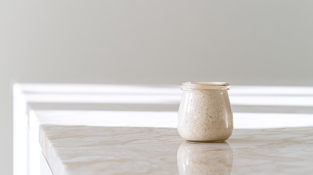 Sourdough starter jar on marble kitchen counter with soft morning light, airy minimalist composition, reflective surface, and spacious neutral background creating calm culinary scene