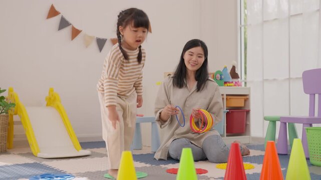 Young Asian girl and adult woman enjoying indoor play with colorful toy in bright playroom, joyful family bonding beside child slide and scattered toys, cheerful activity scene