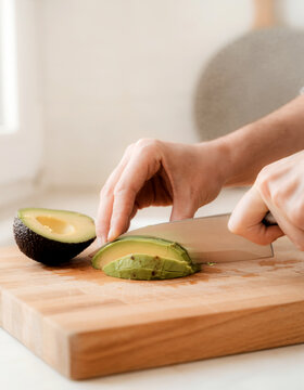 Hands slicing a ripe avocado on a wooden cutting board in a bright kitchen, showcasing fresh healthy food preparation and home cooking