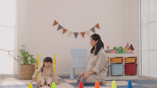 Mother clapping beside toddler during cheerful indoor play, colorful toy activity, festive bunting, family bonding, child development, mother helping daughter stand with support, joyful nursery