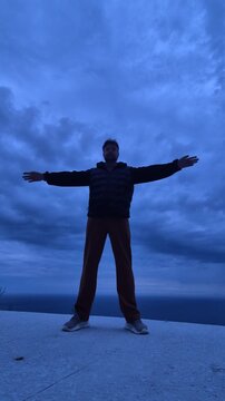 Man stretching standing on a cliff edge under cloudy evening skies by the ocean. Stationary static wide shot.