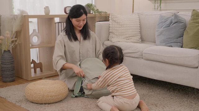 Mother and child packing backpack while Asian parent and toddler play indoors cozy living room, sitting beside fabric basket and sofa, creating warm caring family moment with natural light and home