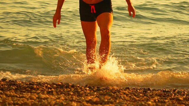 Legs walking, man exits sea onto pebble shore during golden hour sunset light. Static medium shot, low camera angle, focus on lower body.