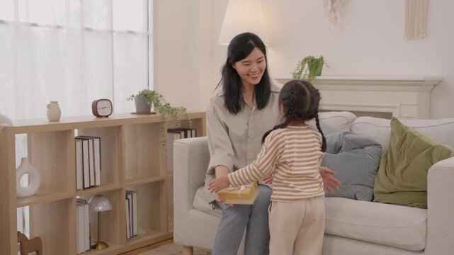 Mother embracing child, joyful family affection celebration living room beside gift box and cozy sofa, Asian mother and daughter sharing warm home moment, smiling woman and child with braid casual