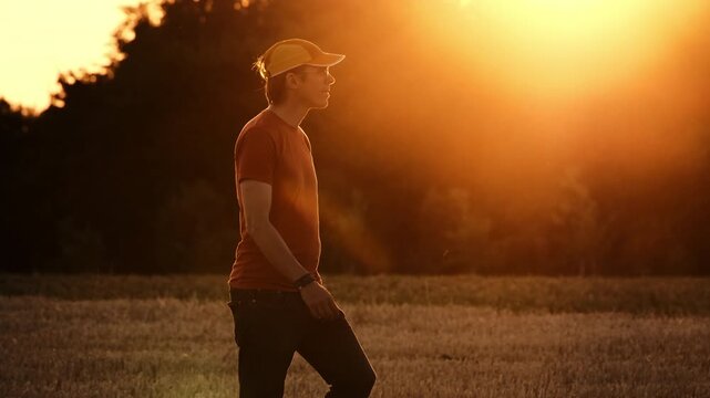 Young man walking on a field at sunset. Smooth hero walk in slow motion, with the sun glowing orange in the background.