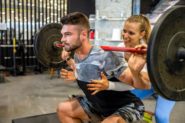 Fototapeta premium Female fitness coach assisting male client with barbell back squat training in modern gym.