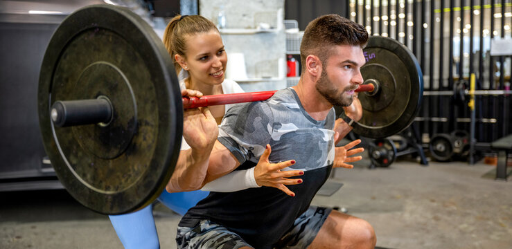 Female personal trainer spotting male athlete during barbell squat exercise in gym.