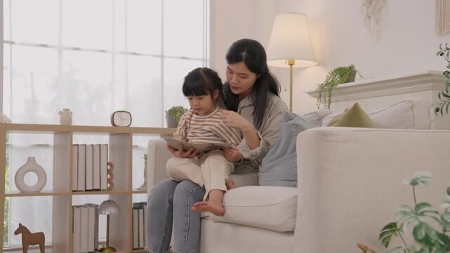 Mother and daughter reading vdo book on sofa in bright living room, tender family moment with child, woman, female parent, warm natural light, home decor, comfort, shared storytelling and connection