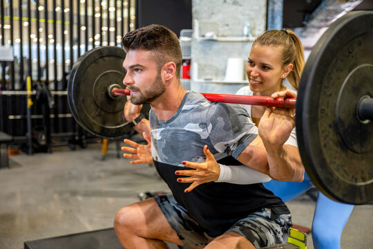 Female fitness coach assisting male client with barbell back squat training in modern gym.