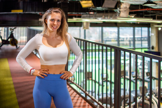 Smiling fit woman standing near balcony railing posing confidently in modern gym.