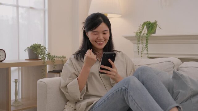 Asian woman smiling using smartphone sofa bright living room, expressing happiness, communication, leisure, calm home atmosphere, female enjoying relaxed modern interior with soft light and houseplant