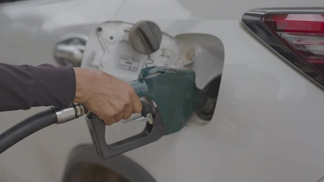 person's hand holding a green fuel pump nozzle, refilling a white car with diesel at a gas station. Concept of automotive transportation, energy cost, and daily driving commute.