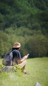 Female hiker using a smartphone while resting in a meadow during a hiking trip with a scenic mountain view, combining outdoor adventure, navigation and digital connectivity.