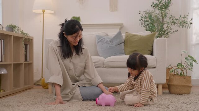 Asian mother and toddler family play with pink toy piggy bank carpet cozy living room home, enjoying warm indoor bonding, savings activity, joyful learning, and meaningful parent child connection