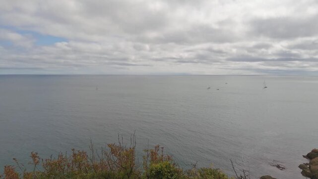 A wide vista overlooking the calm sea, with scattered sailboats visible on the horizon. The shot is taken from the cliffside near St. Catherine's Castle in Fowey, Cornwall.