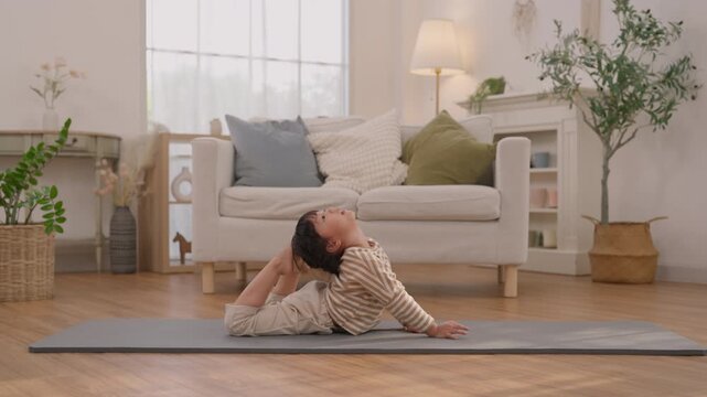 Child stretching on yoga mat in living room, practicing flexibility exercise beside sofa and indoor plant, seated backbend, calm expression, barefoot striped shirt, peaceful indoor wellness scene