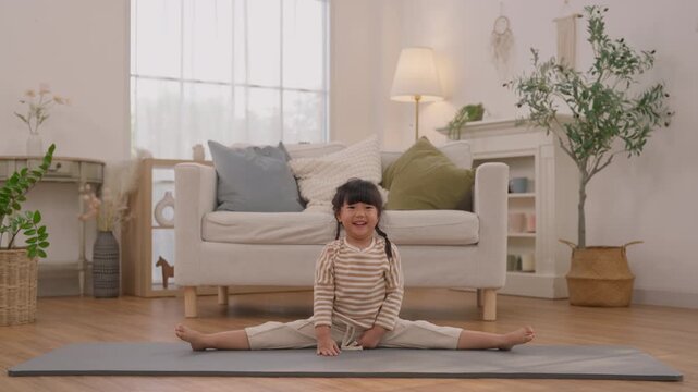Child seated split exercise mat bright living room, smiling with arm extended during playful indoor fitness and yoga practice, joyful stretch beside sofa, houseplant, soft furniture, and natural