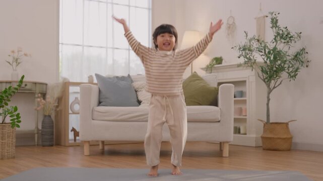 Child Girl Dancing Bright Living Room Home, Smiling Striped Shirt and Beige Pants, Indoor Portrait with Outstretched Arms Near Comfortable Sofa and Indoor Plant, Joyful Energy Asian Family Lifestyle