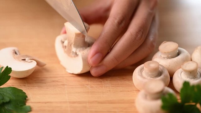 Close-up of woman's hands slicing fresh champignon mushrooms thinly with a knife on a wooden cutting board
