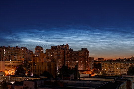 Noctilucent clouds in the twilight sky above residential buildings of a night city.