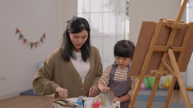 Asian woman and child painting together beside easel in colorful home art studio, creative education activity with vibrant supplies, focused family bonding and joyful artistic expression indoors
