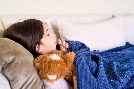 Sick child recovery at home, young girl resting with teddy bear under a blanket in a bright living room
