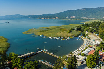 Fototapeta premium Aerial drone view of Struga in North Macedonia with Black Drin River flowing into crystal clear Lake Ohrid, boats, sunny summer landscape, waterfront buildings and scenic city panorama