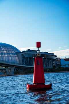 Red maritime navigation buoy on river water with modern highway bridge and glass dome.
