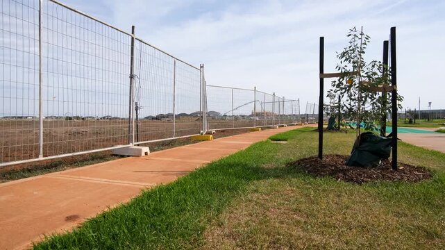 A newly paved footpath along temporary construction fencing adjacent to a new suburban housing development site. Young saplings staked in nature strip, urban growth and land. Melbourne, Australia