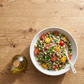 Healthy quinoa salad with cherry tomatoes, chickpeas, cucumber and herbs in white bowl on rustic wooden table with olive oil
