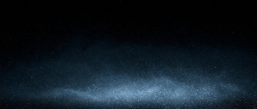 Illuminated blue dust particles and mist rising on dark background. Subtle blue illumination highlights textured dust grains and floating specks. Winter snow