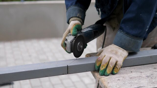 Close-up shows gloved worker using handheld angle grinder on gray metal beam at outdoor construction site, bright sparks scattering from steel surface during focused fabrication task