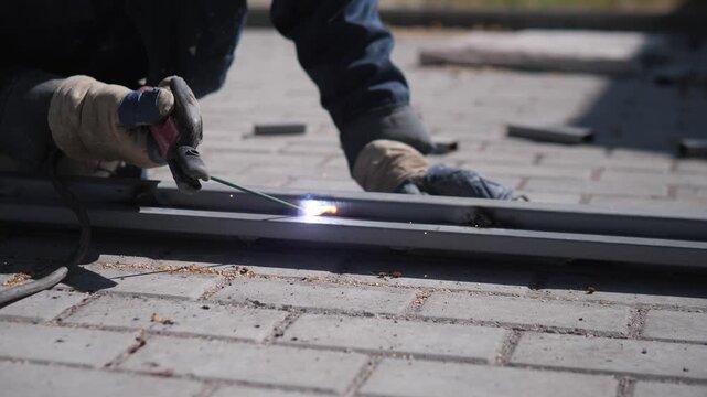 Gloved worker welds long metal rail on brick paved ground, bright arc and drifting sparks showing steady hand control, dark workwear, soft focus outdoor background, muted gray blue tones, rough